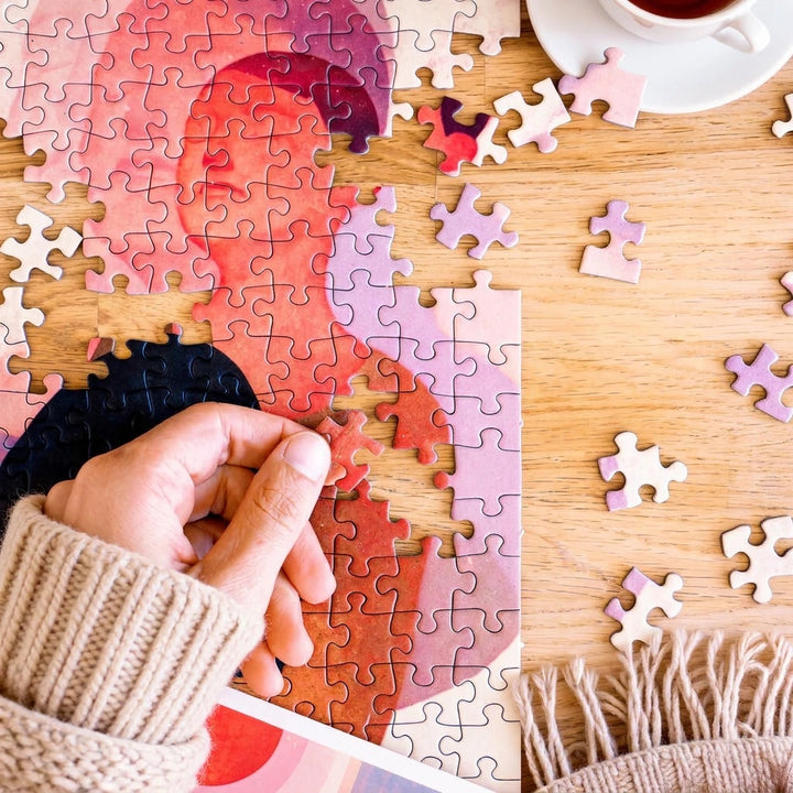 Hands assembling a jigo art puzzle on a wooden table with tea